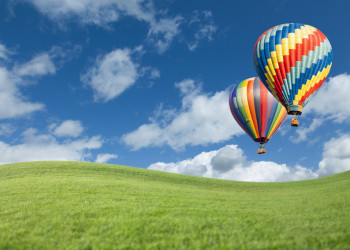 Two Hot Air Balloons Up In The Beautiful Blue Sky With Grass Field Below.